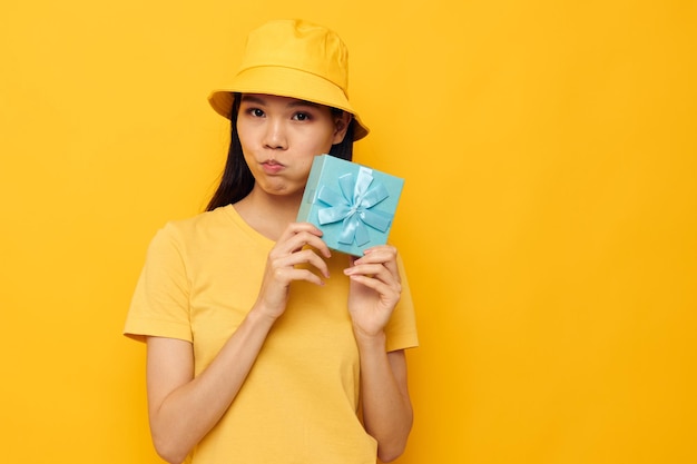 Femme d'apparence asiatique portant un t-shirt jaune et un chapeau avec un cadeau sur fond jaune inaltéré