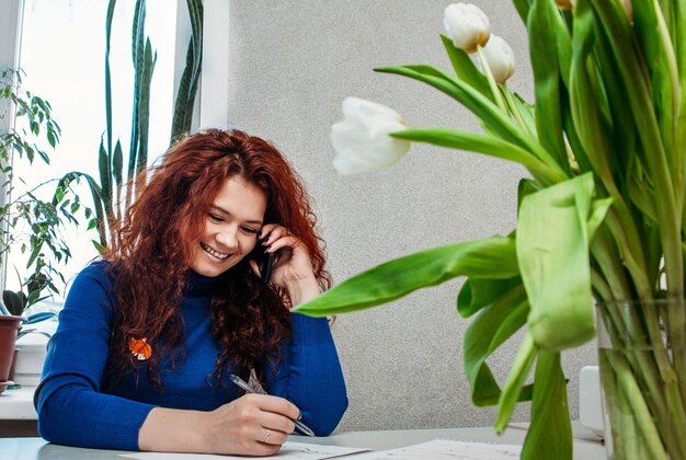 Une femme d'apparence rousse parlant au téléphone en riant et en écrivant quelque chose sur papier