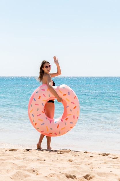 Une femme appelle à nager dans la mer et agite la main. Fille se détendre avec un beignet sur la plage et jouer avec un anneau gonflable. Vacances d'été et concept de vacances.