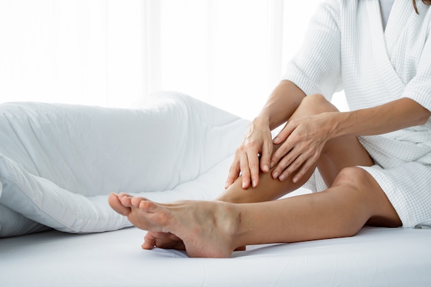 Femme appliquant la crème pour le corps sur sa jambe sur le lit blanc.