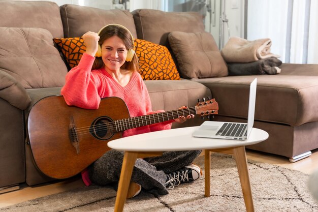 Femme apprenant à jouer de la guitare à la maison avec des cours en ligne sur l'ordinateur en souriant parce qu'elle a bien joué la chanson