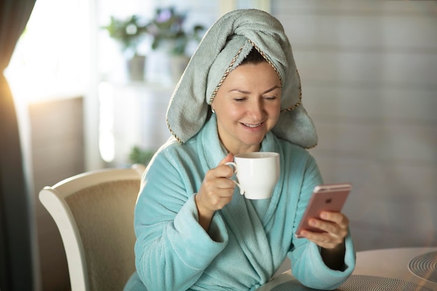 femme après la douche le matin regarde dans le téléphone avec une tasse de café