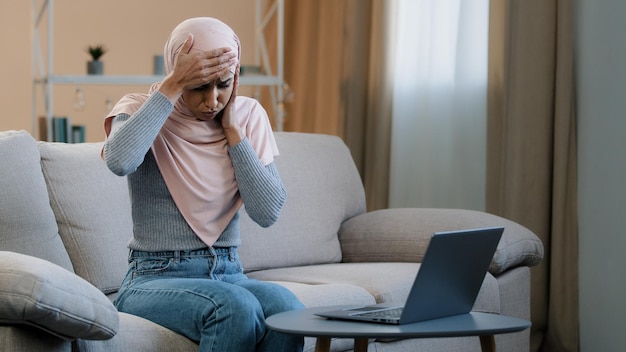 Femme arabe islamique musulmane en hijab rose assis sur un canapé travaillant à l'aide d'un ordinateur portable ressentir le stress des maux de tête