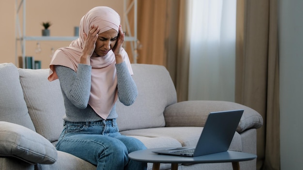 Femme arabe islamique musulmane en hijab rose assis sur un canapé travaillant à l'aide d'un ordinateur portable ressentir le stress des maux de tête