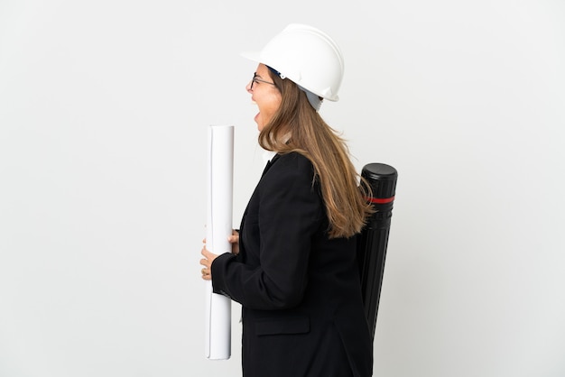 Femme architecte d'âge moyen avec casque et tenant des plans sur fond isolé en riant en position latérale