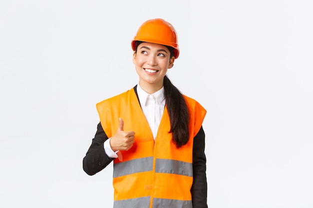 Une femme architecte asiatique souriante et satisfaite, satisfaite des travaux de construction, debout dans un casque de sécurité et une veste réfléchissante, regardant dans le coin supérieur gauche et montrant le pouce levé en signe d'approbation