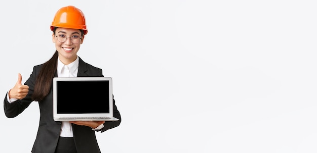 Femme architecte professionnelle souriante responsable de la construction à l'usine montrant un diagramme positif graphique faire le pouce levé en approbation et tenant un ordinateur portable face à l'écran porter un casque de sécurité
