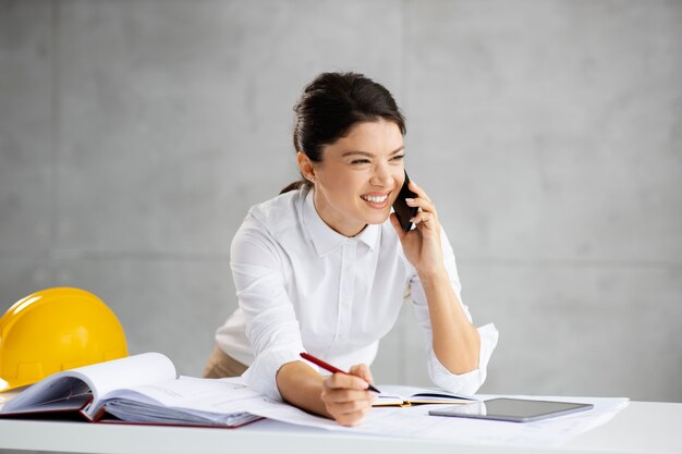 Femme architecte souriante ayant un appel téléphonique avec un client important et notant des informations importantes.