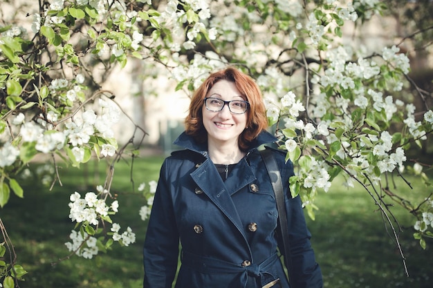 Femme arménienne d'âge moyen confiante dans un trench-coat bleu et des lunettes sous l'arbre en fleurs à la surprise et au sourire