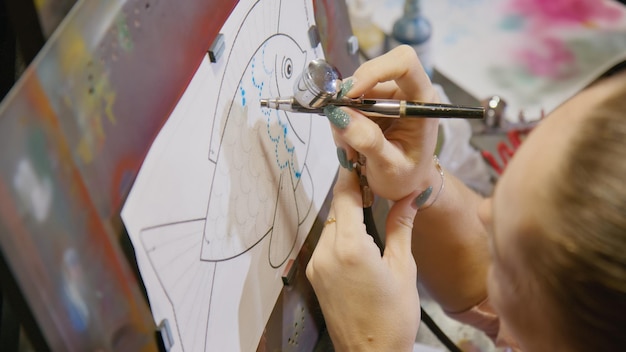 Une femme artiste apprend à peindre à l'aérographe avec du papier de teinture acrylique et un chevalet à l'intérieur