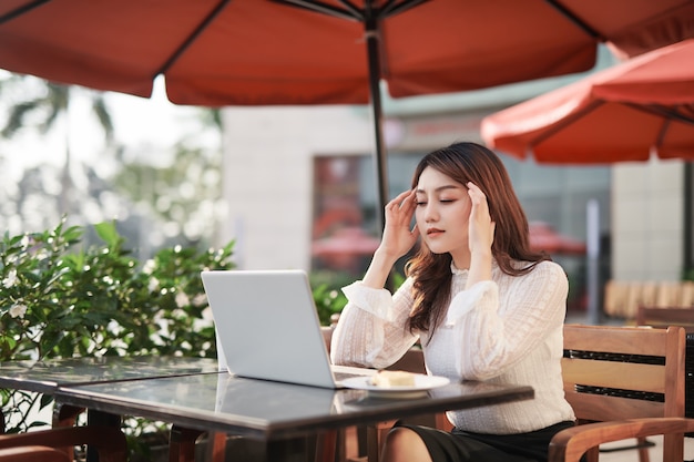 Une femme asiatique adulte travaille sur un ordinateur portable dans un café à l'extérieur de la rue ayant un mal de tête se sentant malade en tenant ses mains sur sa tête