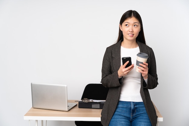 Femme asiatique d'affaires dans son lieu de travail isolé sur fond blanc tenant du café à emporter et un mobile tout en pensant à quelque chose