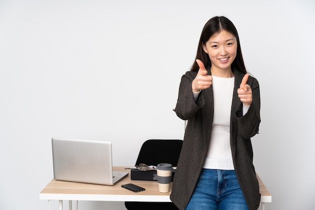 Femme asiatique d'affaires dans son lieu de travail sur un mur blanc pointant vers l'avant et souriant