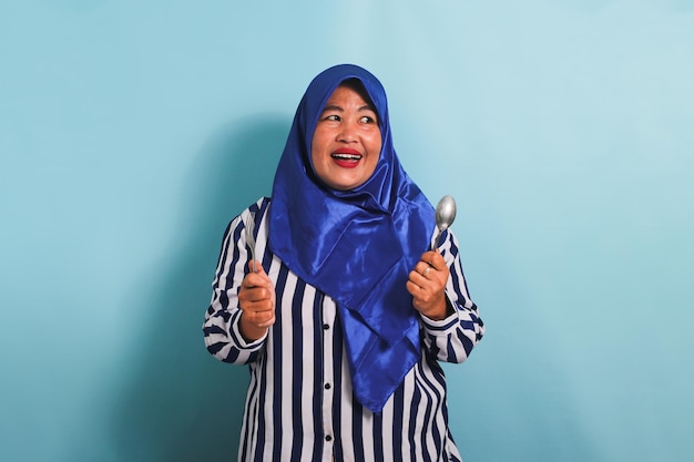 Une femme asiatique affamée d'âge moyen en hijab tient une cuillère et une fourchette isolées sur un fond bleu