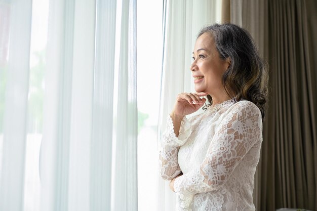 Une femme asiatique d'âge moyen détendue regarde par la fenêtre en rêvant de bonheur dans sa vie