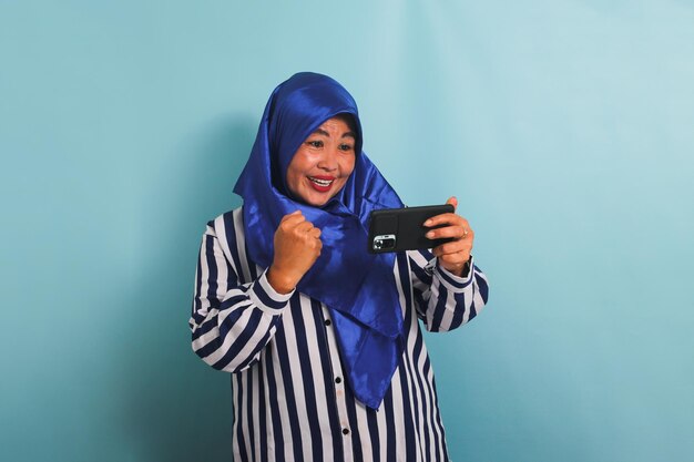 Une femme asiatique d'âge moyen excitée en hijab jouant à un jeu sur son téléphone portable isolée sur un fond bleu
