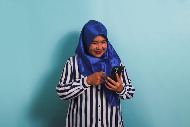 Une femme asiatique d'âge moyen excitée en hijab tient un téléphone portable isolé sur un fond bleu