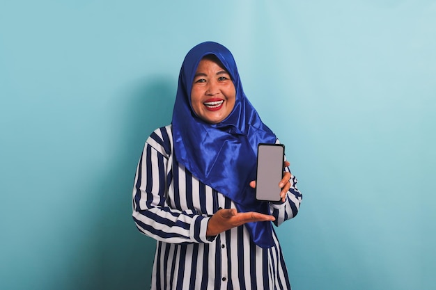 Une femme asiatique d'âge moyen en hijab montre un téléphone isolé sur un fond bleu.