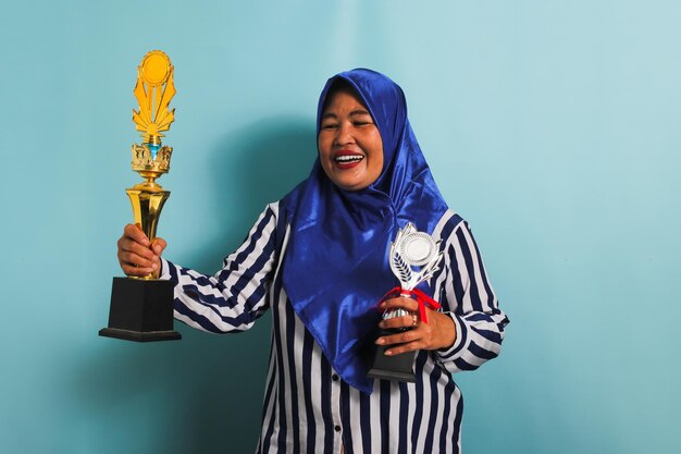 Une femme asiatique d'âge moyen en hijab tient un trophée en or et en argent isolé sur un fond bleu