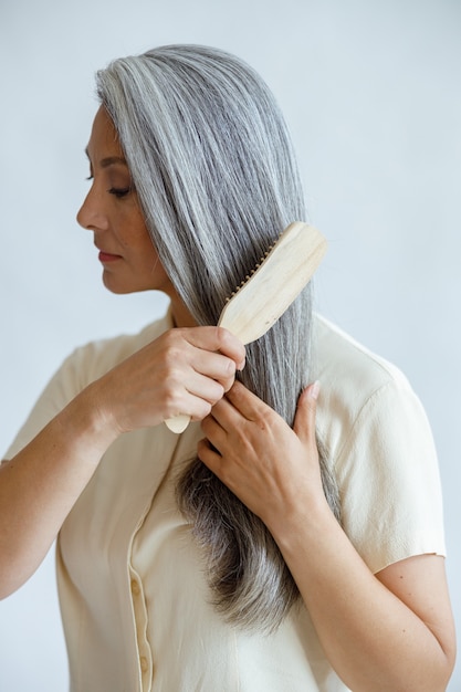 Une femme asiatique d'âge moyen languissante brosse les cheveux blancs naturels sur fond gris clair