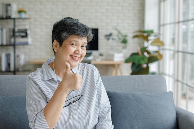 Femme asiatique d'âge moyen regardant la caméra avec le sourire et montrant le geste du pouce vers le haut alors qu'elle était assise sur un canapé dans le salon à la maison