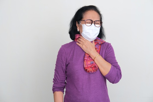 Femme asiatique âgée portant un masque médical touchant sa gorge avec une expression de douleur