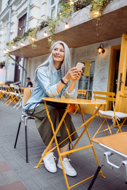 Une femme asiatique âgée réfléchie avec un téléphone est assise à une petite table sur une terrasse de café en plein air