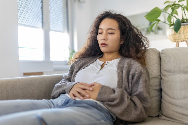 Femme asiatique allongée sur un canapé à la recherche de malades dans le salon Belle jeune femme allongée sur le lit et se tenant la main sur le ventre