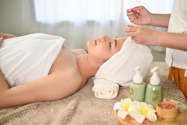 Femme asiatique allongée sur une table de massage recevant un masque d'argile de traitement facial par un travailleur du spa