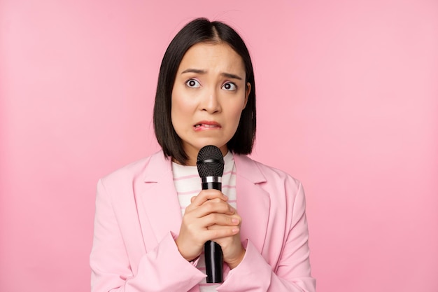 Une femme asiatique anxieuse en costume parlant en public prononçant un discours avec microphone lors d'une conférence ayant l'air effrayée debout sur fond rose