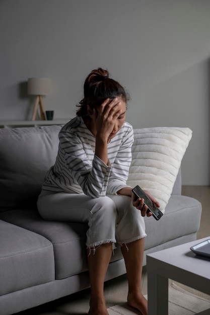 Photo femme asiatique assise sur le canapé tenant le téléphone se sentant déçue triste bouleversée femme souffrant de problèmes de santé mentale