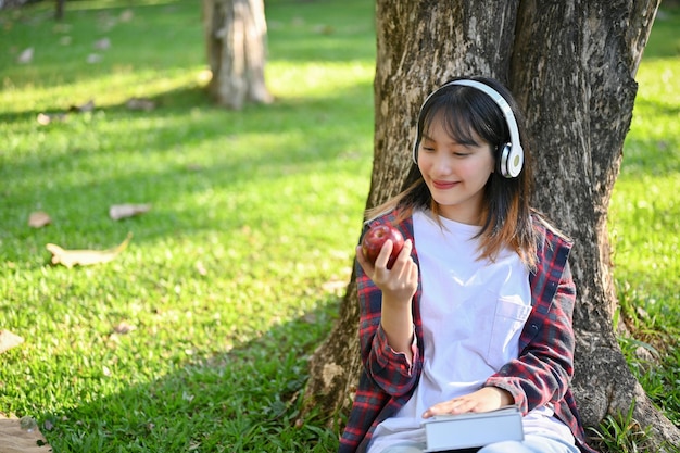 Femme asiatique assise sous l'arbre écoutant de la musique à travers ses écouteurs et mangeant une pomme