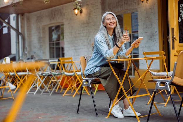 Une femme asiatique aux cheveux de proie dans des vêtements décontractés tient un verre et un smartphone à table à l'extérieur