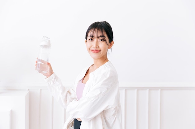 Femme asiatique aux longs cheveux noirs ligotée portant des vêtements de sport et une veste de sport blanche Heureux souriant à la caméra et tenant une bouteille d'eau debout sur un fond blanc Image avec espace de copie