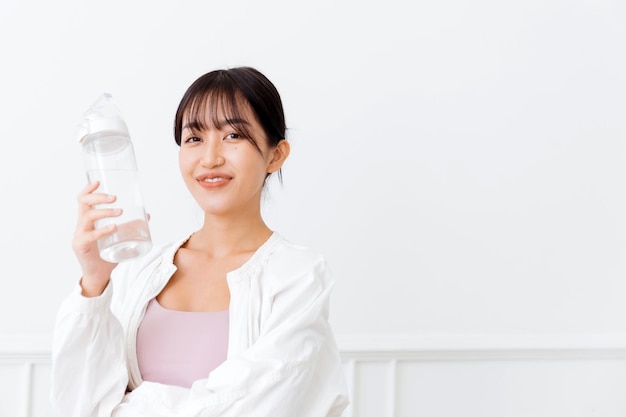 Femme asiatique aux longs cheveux noirs ligotée portant des vêtements de sport et une veste de sport blanche Heureux souriant à la caméra et tenant une bouteille d'eau debout sur un fond blanc Image avec espace de copie