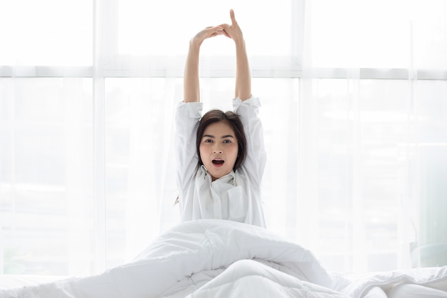 Femme asiatique Belle jeune femme souriante assise et dormant dans un lit blanc et s'étirant le matin dans la chambre après s'être réveillée dans son lit complètement reposée