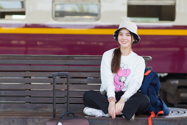 Une femme asiatique en blanc à manches longues dans un chapeau est assise en souriant sur une chaise avec son sac à la plate-forme du train