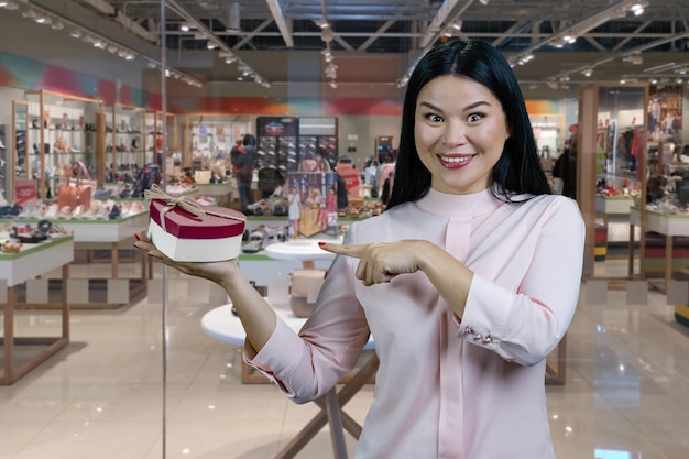 Femme asiatique brune pointant sur l'intérieur de la boutique de chaussures en forme de coeur en arrière-plan