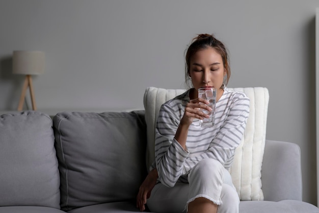 Femme asiatique buvant de l'eau froide tout en étant assise sur le canapé à la maison Femme buvant de l'eau propre santé