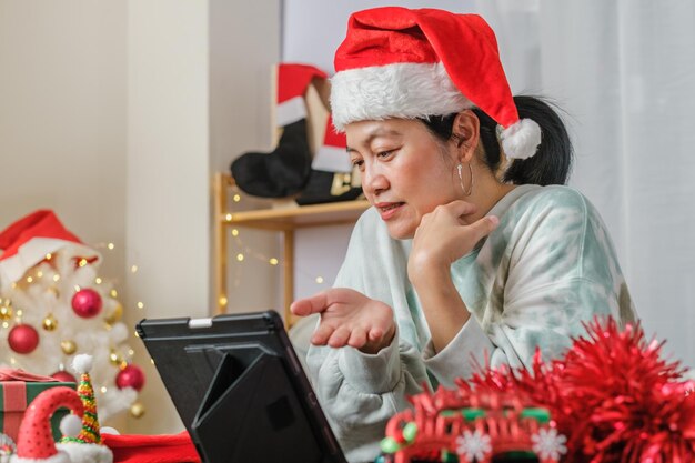 Une femme asiatique célèbre le nouvel an et la fête de noël avec des amis d'appel vidéo sur tablette à la maison