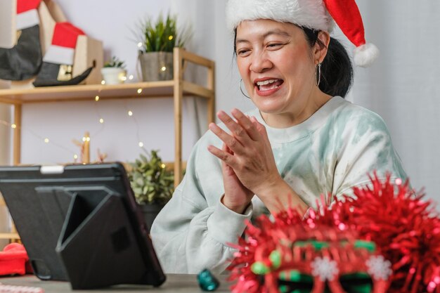 Une femme asiatique célèbre le nouvel an et la fête de noël avec des amis d'appel vidéo sur tablette à la maison