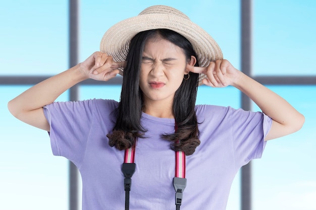 Une femme asiatique avec un chapeau et un appareil photo a fermé ses oreilles avec un doigt à l'intérieur du bâtiment avec un fond de ciel bleu