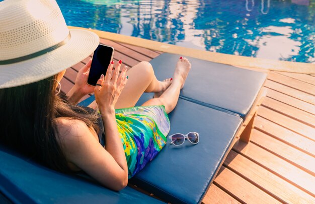 Femme asiatique avec chapeau et maillot de bain assis sur un transat au bord de la piscine et à l'aide de smartphone en vacances d'été au bord de la piscine.