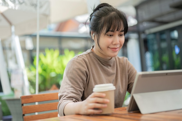 Une femme asiatique charmante positive portant des écouteurs et travaillant sur sa tablette numérique dans un café