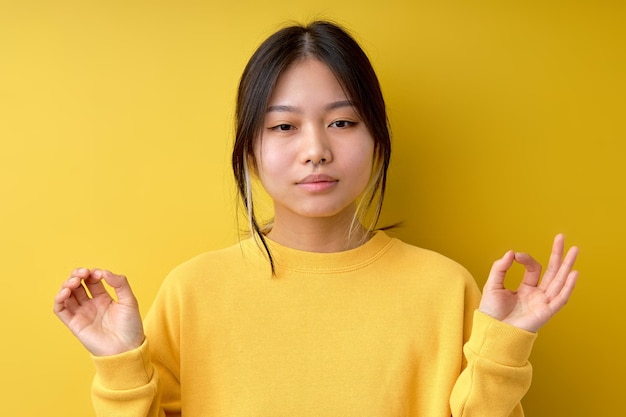 Une femme asiatique ou chinoise calme et confiante fait un geste zen nirvana médite en regardant la caméra pratique le yoga une femme brune porte un pull jaune décontracté posant isolé sur fond jaune