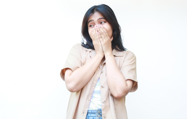 femme asiatique choquée couvrant sa bouche portant des vêtements bruns décontractés isolés sur fond blanc