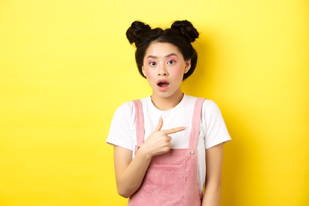Femme asiatique choquée avec maquillage glamour, bouche ouverte et pointant à droite sur le logo, montrant la publicité, debout sur fond jaune