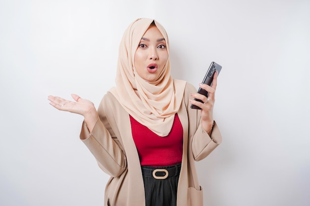 Femme asiatique choquée portant le hijab pointant vers l'espace de copie à côté d'elle tout en tenant son téléphone isolé sur fond blanc