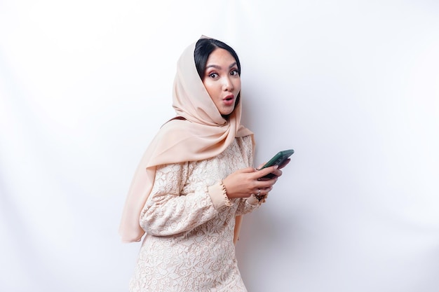 Femme asiatique choquée portant le hijab tenant son téléphone isolé sur fond blanc