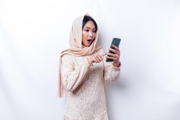 Femme asiatique choquée portant le hijab tenant son téléphone isolé sur fond blanc